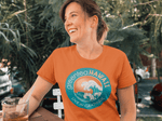 smiling woman sitting outside with big green leaves behind her wearing an orange t-shirt featuring the greenteaHAWAII logo holding a cup of greenteaHAWAII